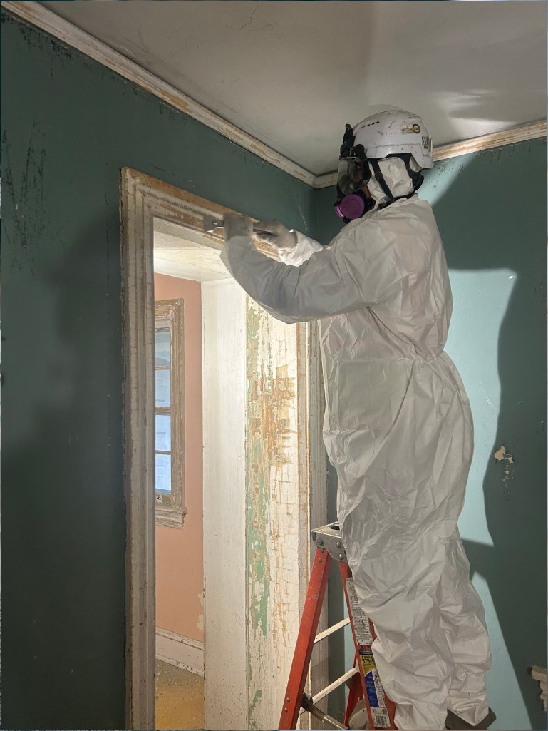 RETRO crew member in a protective suit, hard hat, and respirator removing lead-based paint from historic door trim