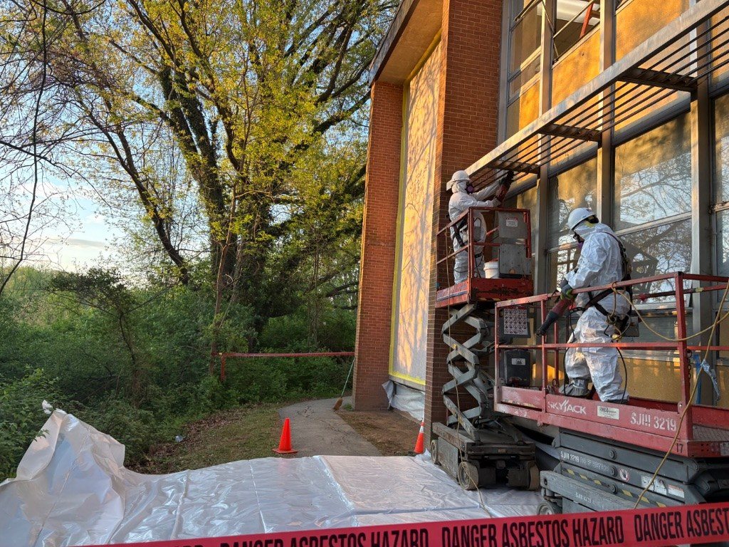 RETRO crew in protective suits and respirators working from scissor lifts on a brick and glass façade, with poly sheeting on the ground