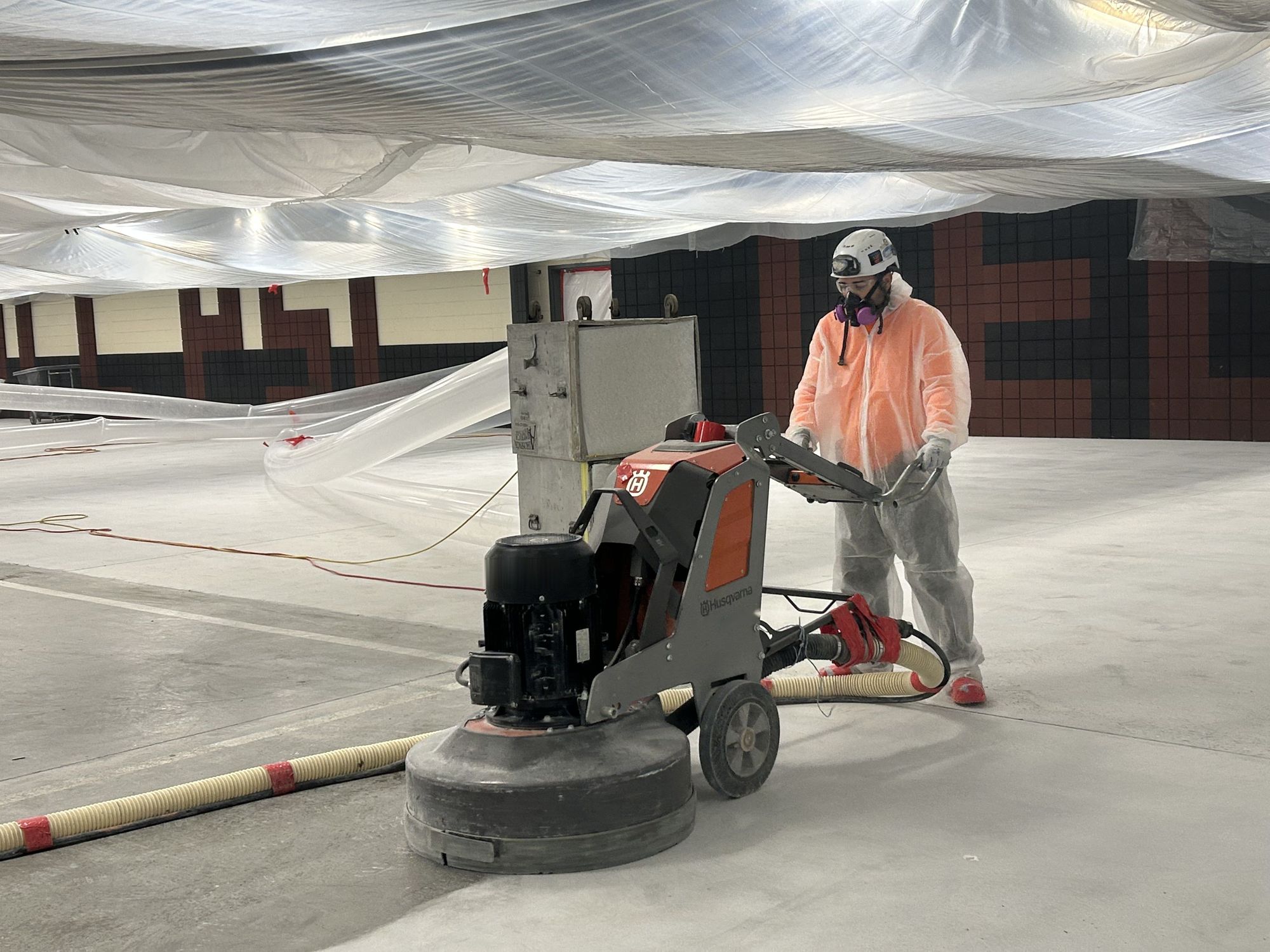 RETRO crew member in a protective suit, hard hat, and respirator running a floor grinder with HEPA dust collection, a negative air machine behind him