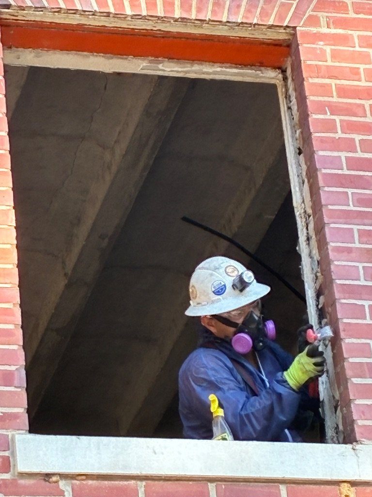RETRO crew member in a protective suit, hard hat, and respirator working at a window opening in a brick wall