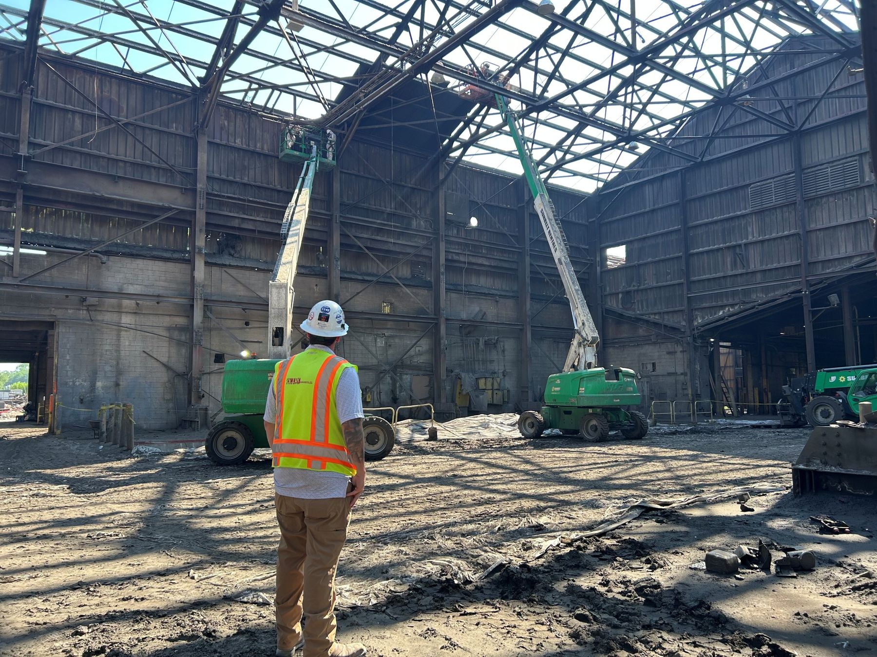 RETRO site lead in a hard hat and RETRO safety vest watching crews on two boom lifts work at the roof steel of a large industrial building