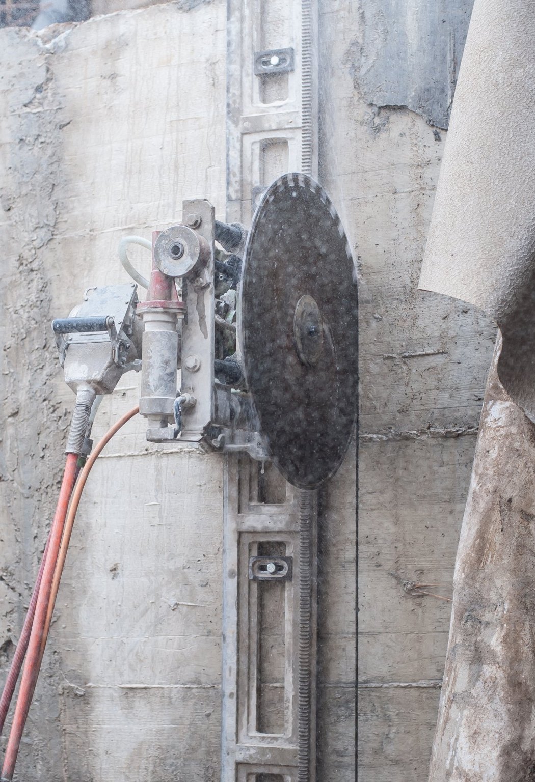 Track-mounted wall saw cutting a reinforced concrete wall, with water spraying from the diamond blade